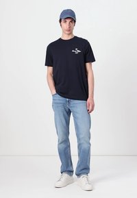 Marineblaue Baumwoll-T-Shirt mit weißem grafischen Logo, helle blaue Jeans und weiße Sneakers. Das Modell trägt eine hellblaue Kappe.