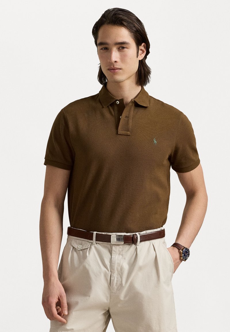 Cămașă polo maro cu mâneci scurte și guler, având un mic emblemă verde. Combinată cu pantaloni de culoare deschisă și o curea maronie.