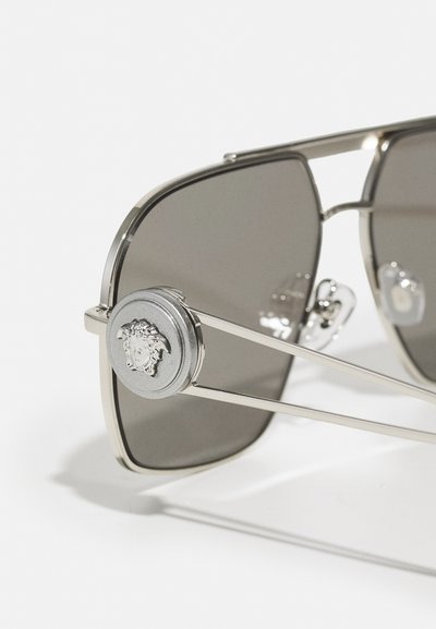 Ochelari de soare cu o ramă din metal argintiu, lentile gri și un accent rotund care prezintă un logo detaliat. Design unghiular și sprijini subțiri.