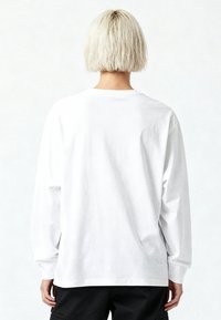 Persona con capelli corti e decolorati, vista di spalle, indossa una maglia bianca a maniche lunghe e pantaloni neri su uno sfondo bianco.