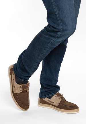 Persona che indossa jeans blu scuro e scarpe da barca in camoscio marrone con dettagli beige, in piedi con un piede leggermente sollevato.
