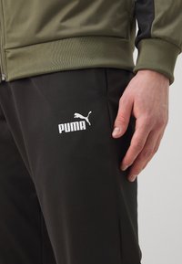 Maslinasto zelena zip-up jakna s tamnim naglascima, uparena s crnim hlačama koje imaju bijeli PUMA logotip. Glatka, sportska tkanina s prilagođenim dizajnom.
