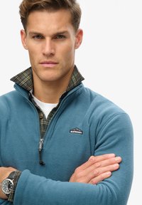 Sudadera de polar azul con cuello con cremallera, que cuenta con un forro de cuadros verde y un pequeño logo en el lado izquierdo del pecho. Textura suave y corte casual.