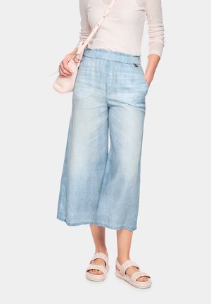 Frau trägt hellblaue, weit geschnittene, gekürzte Jeans, blassrosa Langarmtop, passende Umhängetasche und blassrosa Sandalen.