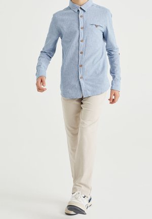 Bambino che cammina indossando una camicia azzurra con bottoni, pantaloni beige e scarpe da ginnastica bianche con dettagli blu navy su sfondo bianco.