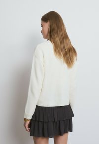 Cremefarbener Strickpullover mit einem gerippten Saum, mit abgesenkten Schulternähten, einem darunter liegenden, mehrlagigen grauen Kleid mit Rüschen sowie sichtbar braunem Haar.