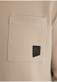 Beige texturovaná látka se čtvercovou náprsní kapsou, na které je malá černá nášivka s oranžovým geometrickým vzorem.