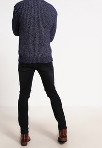 Marinblå chunky stickad tröja med en texturerad design, tillsammans med smal passform svarta byxor och bruna läderstövlar. Vy från baksidan.