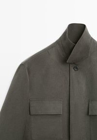 Olivgrüne Jacke mit Stehkragen, Knopfverschluss und zwei Klapp-Brusttaschen vor weißem Hintergrund.