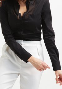 Czarna koszula zapinana na guziki z przezroczystymi detalami i dwiema kieszeniami na piersiach, zestawiona z białymi spodniami o dopasowanym kroju z gładkim pasem.