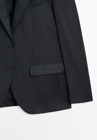 Blazer nero su misura realizzato in tessuto testurizzato, caratterizzato da due bottoni frontali, una tasca sul petto e una finitura pulita sui bordi.