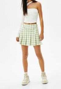 Grön och vit rutmönstrad plisserad kjol med en silverkedjedetalj, kombinerad med en vit ribbad topp med en axel och beige sneakers.