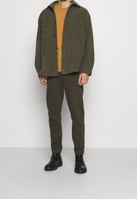 Olivově zelená strukturovaná košile-jacket přes hořčicově žluté tričko, v kombinaci s tmavě zelenými kalhotami a černými kotníkovými botami.