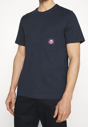 Un homme porte un t-shirt bleu marine foncé à manches courtes avec un petit patch logo rectangulaire au-dessus de la poche poitrine gauche, associé à un pantalon noir.