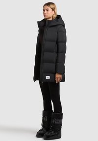 Fekete steppelt dzseki cipzárral, magas gallérral és fedett varratokkal. Fekete szigetelt csizmával párosítva, mely fűzővel és logó díszítéssel rendelkezik.