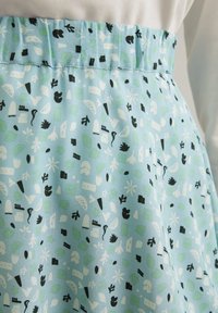 Jupe en tissu bleu clair avec une taille élastique ; présente un motif abstrait en blanc, noir et vert, incluant des feuilles et des formes géométriques.