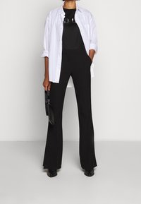 Camisa blanca de botones sobre una camiseta negra con letras brillantes, combinada con pantalones de campana negros y zapatos negros, sosteniendo un bolso negro.