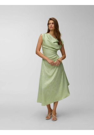 Femme portant une robe midi sans manches vert clair avec un décolleté drapé, des sandales et des bracelets, debout sur un fond clair uni.