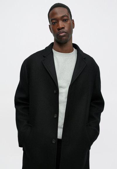 Fekete gyapjú kabát, lekerekített gallérral, gombos záródással és első zsebekkel, világosszürke kereknyakú pulóver felett. Sima textúrájú és testhez simuló fazonú.