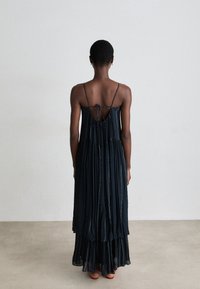 Vestido maxi plissado em azul-marinho com alças finas, detalhe nas costas abertas, design em camadas e acentos texturizados contrastantes. Barra rotativa perto dos tornozelos.