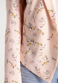 Veste douce rose à motif floral avec un tissu texturé et des zips métalliques à l'avant. Design ajusté avec un ourlet incliné.