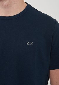 T-shirt in cotone blu navy con scollatura rotonda, maniche corte e un piccolo logo geometrico ricamato in bianco sul lato sinistro del petto.