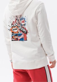 Felpa leggera beige con una grafica in stile fumetto sulla schiena, che presenta testi come "POW!" e "BOOM!" in colori vivaci e forme dinamiche.