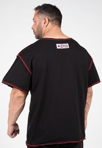 Black oversized t-shirt with red stitching detail. Features a tag on the back and raw edges on sleeves and hem. Cotton material.