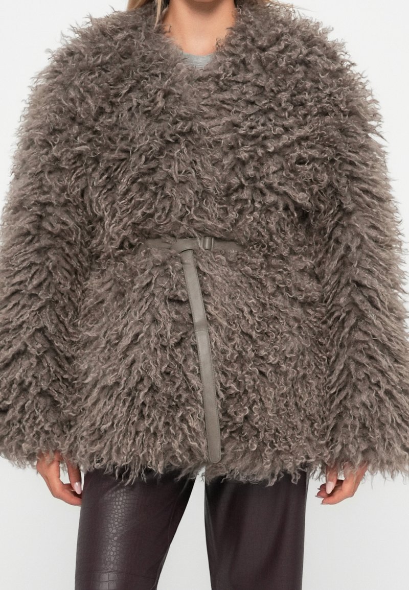 Femme portant un manteau volumineux en fausse fourrure marron bouclée avec une fine ceinture, associé à un pantalon texturé foncé.