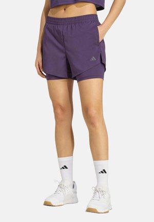 Personne portant un short de sport violet superposé, des chaussettes blanches mi-mollet avec un logo noir, et des chaussures de sport blanches avec semelles en gomme.