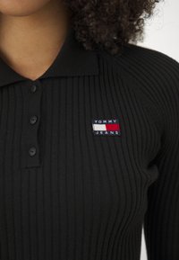 Polo negro de canalé que presenta una tapeta con botones y un parche rectangular con elementos en rojo, blanco y azul.