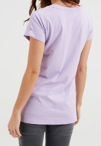 Kvinna i en lavendel t-shirt med uppvikta ärmar och mörkgrå jeans, sedd bakifrån mot en enfärgad vit bakgrund.