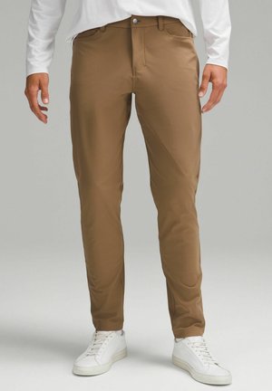 Mann trägt braune Slim-Fit-Hosen und weiße Sneakers und steht vor einem neutralen grauen Hintergrund.