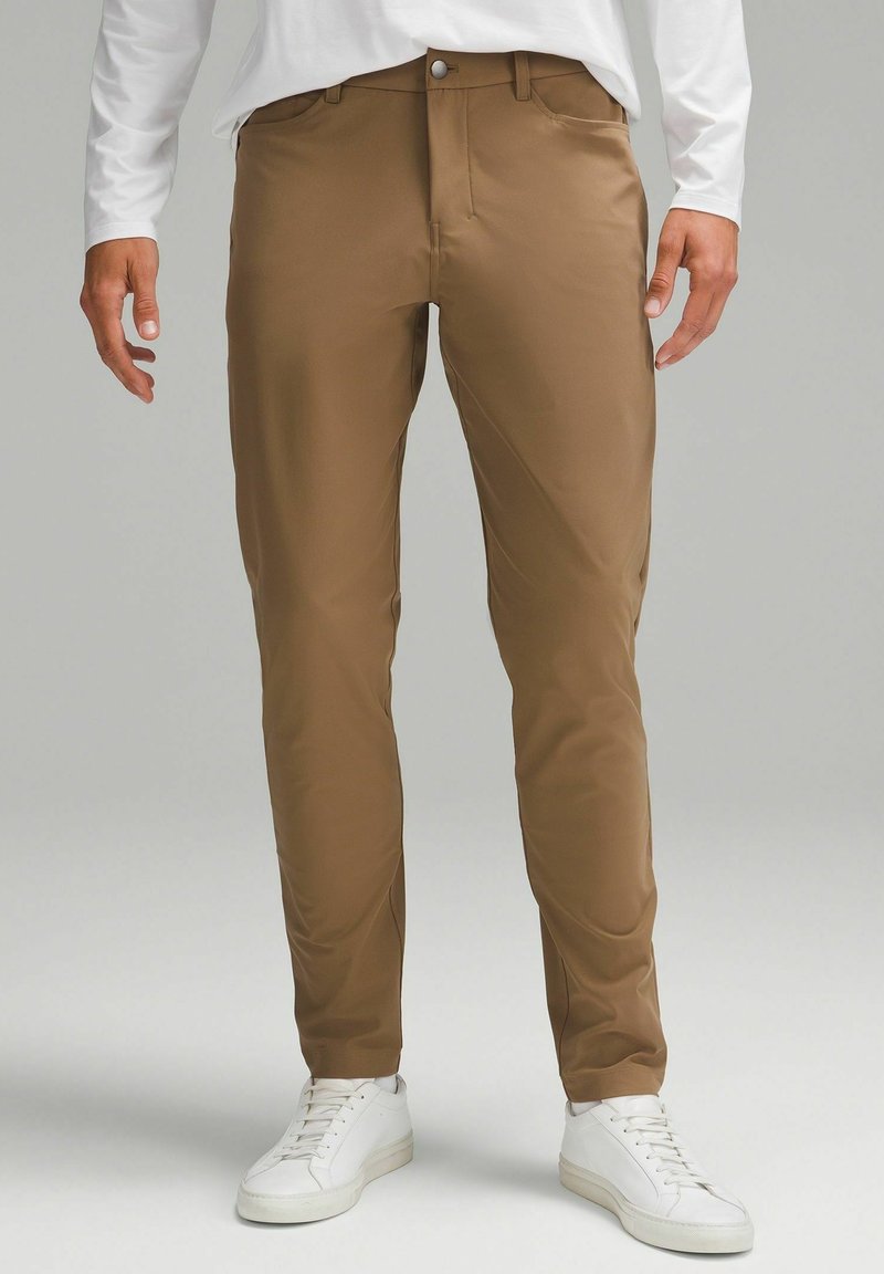 Homme portant un pantalon marron coupe slim et des baskets blanches, debout devant un fond gris neutre.