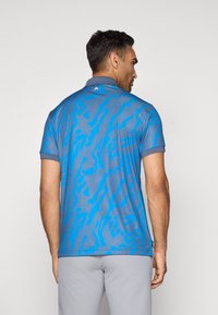 Homme portant un polo bleu à manches courtes avec des motifs abstraits bleu foncé, associé à un pantalon gris clair, vu de dos.
