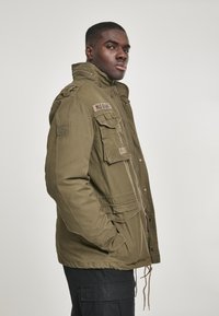 Olive green military-style jacket with a high collar, multiple front pockets, zipper, and adjustable drawstring at the waist.