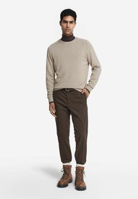 Beige Strickpullover, braune Hose und braune Schnürstiefel. Der Pullover hat gerippte Bündchen, die Hose ist schmal geschnitten. Weiche Texturen und neutrale Farben.