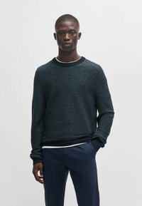 Dunkel strukturierter Pullover mit Rundhalsausschnitt, gerippten Bündchen und Saum. Getragen über einem weißen Hemd, kombiniert mit navyblauen Hosen.