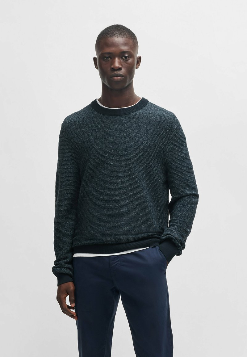 Dunkel strukturierter Pullover mit Rundhalsausschnitt, gerippten Bündchen und Saum. Getragen über einem weißen Hemd, kombiniert mit navyblauen Hosen.