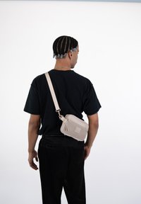 Borsa a tracolla beige in materiale testurizzato, forma rettangolare, chiusura con zip e tracolla regolabile; esposta su una persona che indossa una maglietta nera.