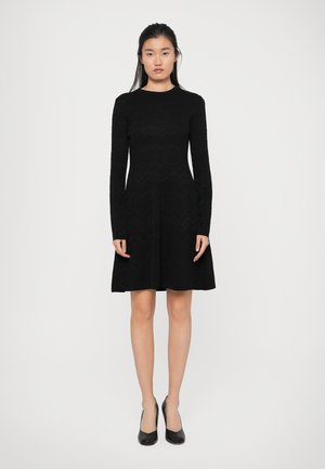 Robe noire à manches longues, motif en chevrons texturé, corsage ajusté, jupe évasée, longueur genoux, associée à des chaussures à talons hauts noires.