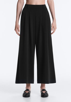 Žena s čiernymi nohavicami s vysokým pásom, širokými nohavicami a záhybmi, v kombinácii s čiernymi slip-on topánkami, stojaca pred jednoduchým pozadím.