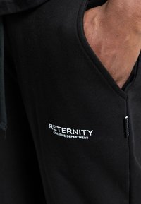 Black jogger pants featuring a side pocket, with "RETERNITY CREATIVE DEPARTMENT" in white text, made from a smooth cotton fabric.