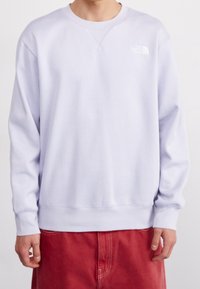 Osoba v světle fialovém crewneck svetru s logem The North Face na prsou a červených kalhotách, stojící na jednobarevném pozadí.