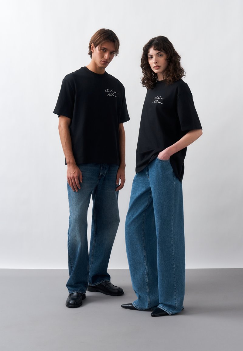 Jeune homme et femme portant des t-shirts noirs Calvin Klein et des jeans bleus, debout devant un fond clair uni.
