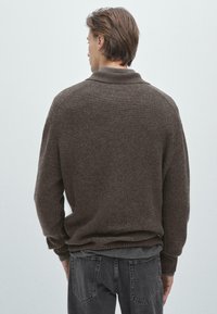 Pull en maille côtelée marron avec col montant, épaules tombantes, et manches longues, associé à un jean gris foncé avec une coupe décontractée.