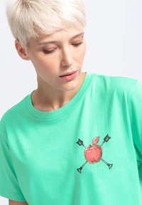 Mintgroene katoenen t-shirt met een centraal grafisch ontwerp van een rode appel gekruist met pijlen, ronde halslijn en korte mouwen.