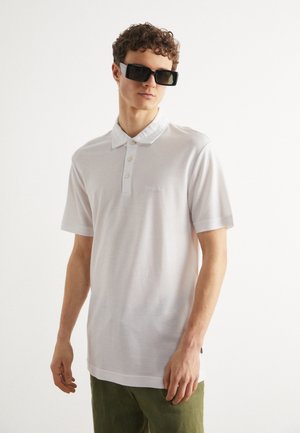Camisa polo blanca de manga corta con cuello puntiagudo, que presenta tres botones y un pequeño logo en el pecho; se lleva con gafas de sol negras.