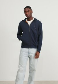 Marine blauwe zip-up hoodie van zacht materiaal, met een contrasterende oranje binnenkant, voorzakken en een relaxte pasvorm. Gecombineerd met lichte jeans.