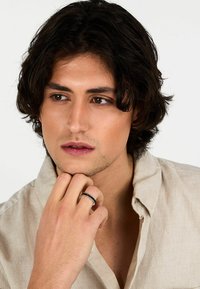 Anello d'argento con un'intarsio nero. Chi lo indossa ha capelli castani ondulati di media lunghezza e una camicia di lino chiara. La pelle appare chiara e liscia.